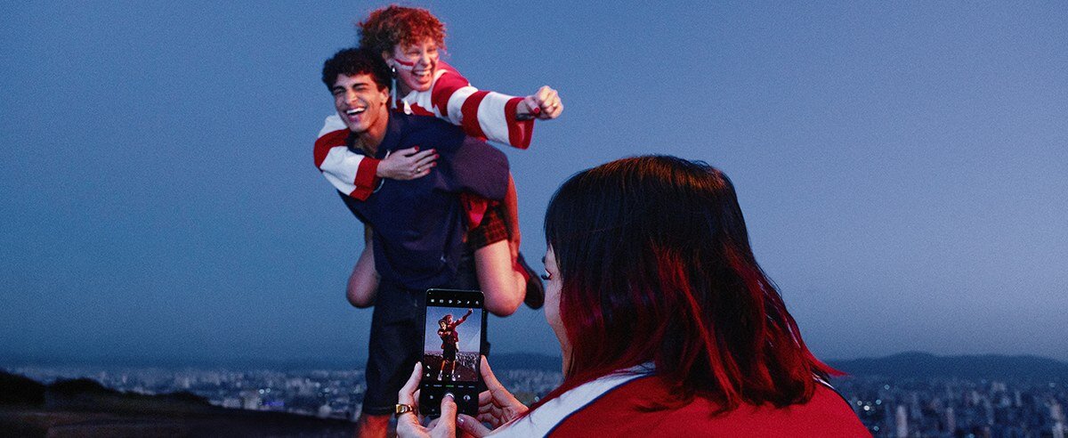 Osoba robi zdjęcie dwóm innym osobom, z których jedna siedzi na plecach drugiej. W tle widać panoramę miasta o zmierzchu.