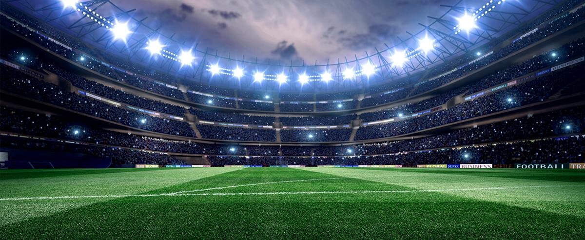 Obraz przedstawia oświetlony stadion piłkarski z zieloną murawą i trybunami w tle. Na trybunach widoczne są reklamy, ale nie można odczytać ich treści.