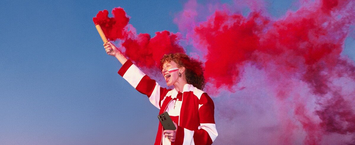 Osoba ubrana w biało-czerwony strój trzyma telefon i czerwoną racę dymną, z której unosi się dym na tle nieba.