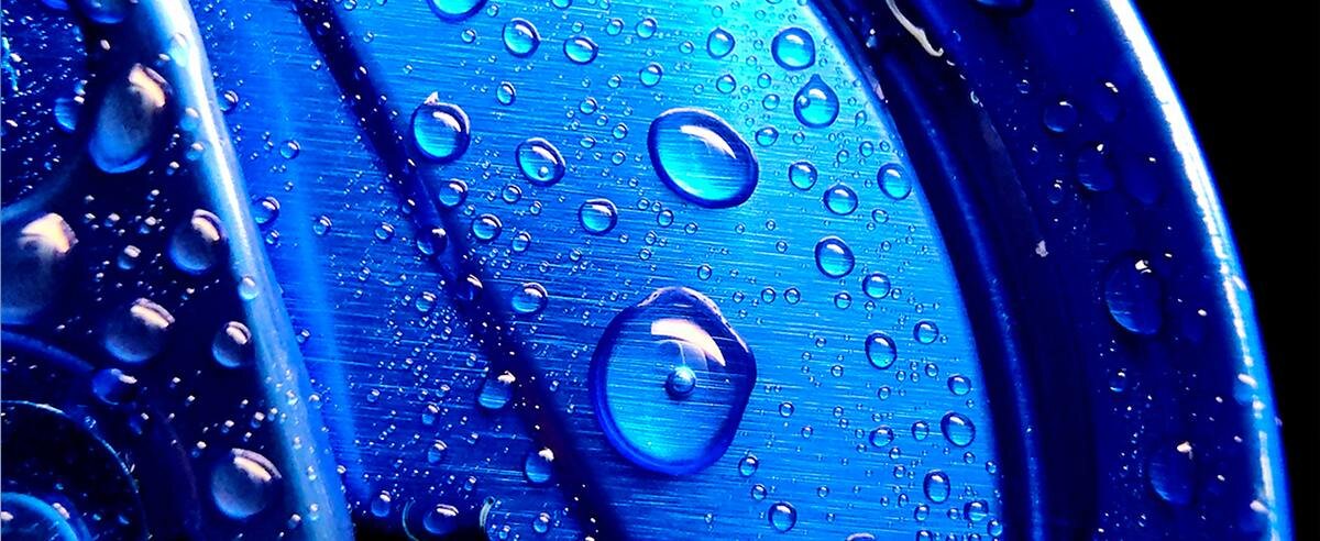 Obraz przedstawia zbliżenie na niebieską powierzchnię z metalicznym połyskiem, pokrytą kroplami wody. Krople mają różne rozmiary i tworzą efekt trójwymiarowości.