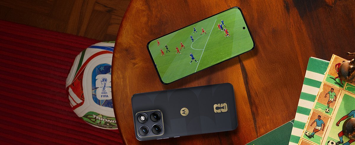 Na drewnianym stole leży smartfon z włączonym ekranem, na którym widać mecz piłki nożnej. Obok znajduje się piłka z logo FIFA i fragment planszy z figurkami piłkarzy.