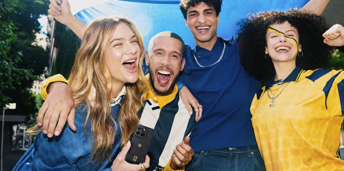 Cztery osoby uśmiechają się i pozują do zdjęcia, jedna z nich trzyma smartfon. W tle widoczne są drzewa i fragment budynku.