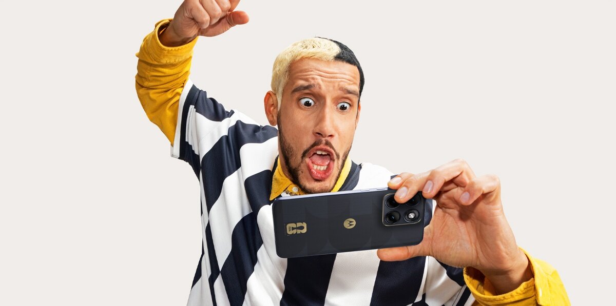 Osoba trzyma smartfon z logo MOTOROLA, ubrana w koszulkę w czarno-białe pasy. Na obudowie telefonu widoczne jest logo FIFA World Cup 26™.
