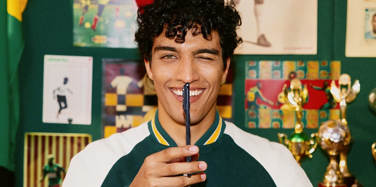 Osoba trzymająca smartfon przed twarzą, w tle widoczne są trofea i plakaty sportowe.