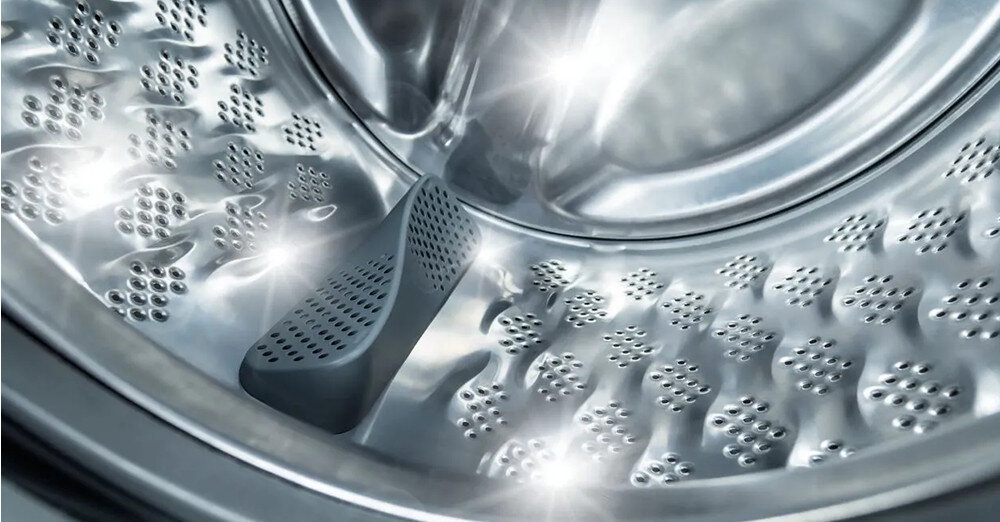 Wnętrze bębna pralki z metaliczną powierzchnią i licznymi otworami. Widoczne są refleksy świetlne na powierzchni metalu.