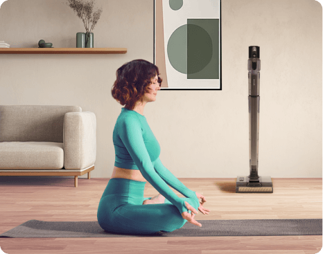 Wnętrze pokoju z sofą, półką z dekoracjami i obrazem na ścianie. Na podłodze stoi odkurzacz pionowy.