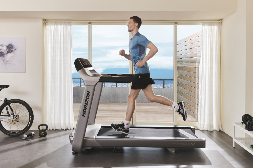 Mężczyzna biegnie na bieżni elektrycznej z widocznym logo \'HORIZON\'. W tle znajduje się rower, hantle i duże okno z widokiem na zewnątrz.