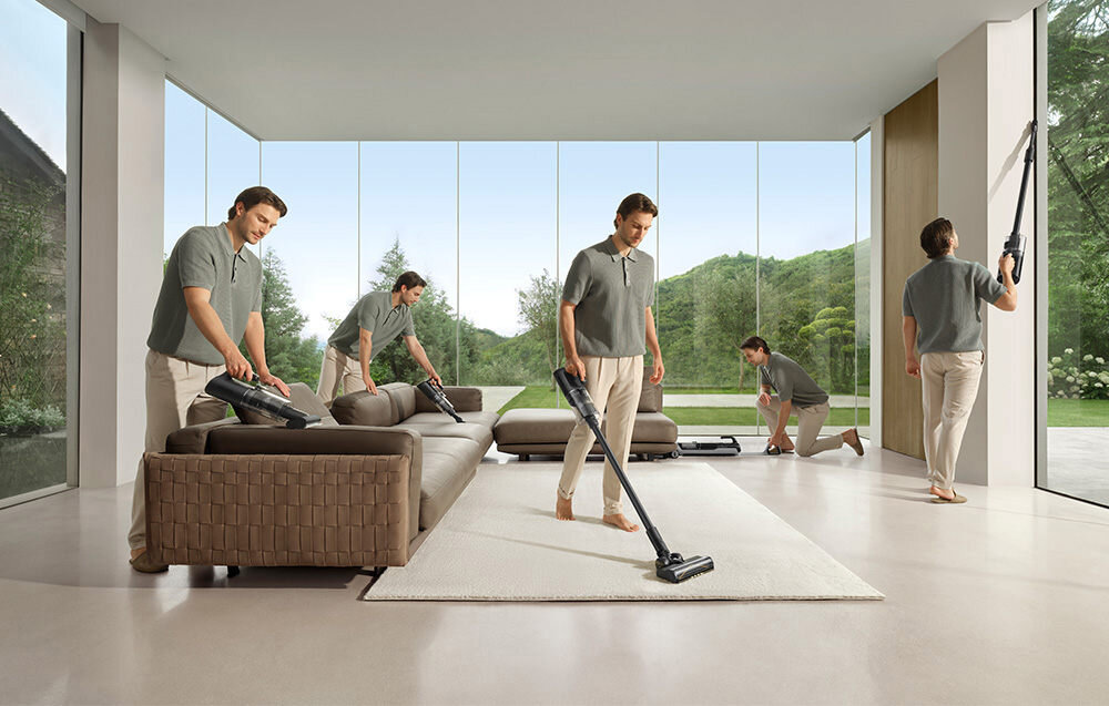 W przestronnym, nowoczesnym salonie z dużymi oknami, kilka osób używa odkurzacza mopującego do czyszczenia różnych powierzchni, w tym dywanu i ścian. W tle widoczny jest zielony krajobraz za oknami.