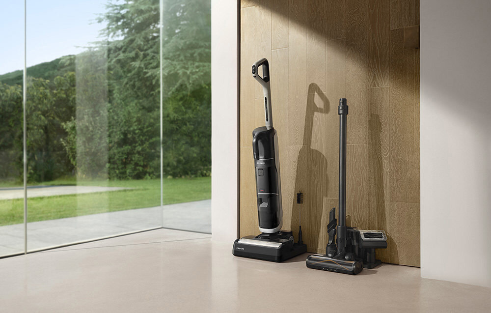 Na zdjęciu widać odkurzacz mopujący stojący na podłodze przy ścianie z drewnianymi panelami. W tle znajduje się duże okno z widokiem na zieleń.