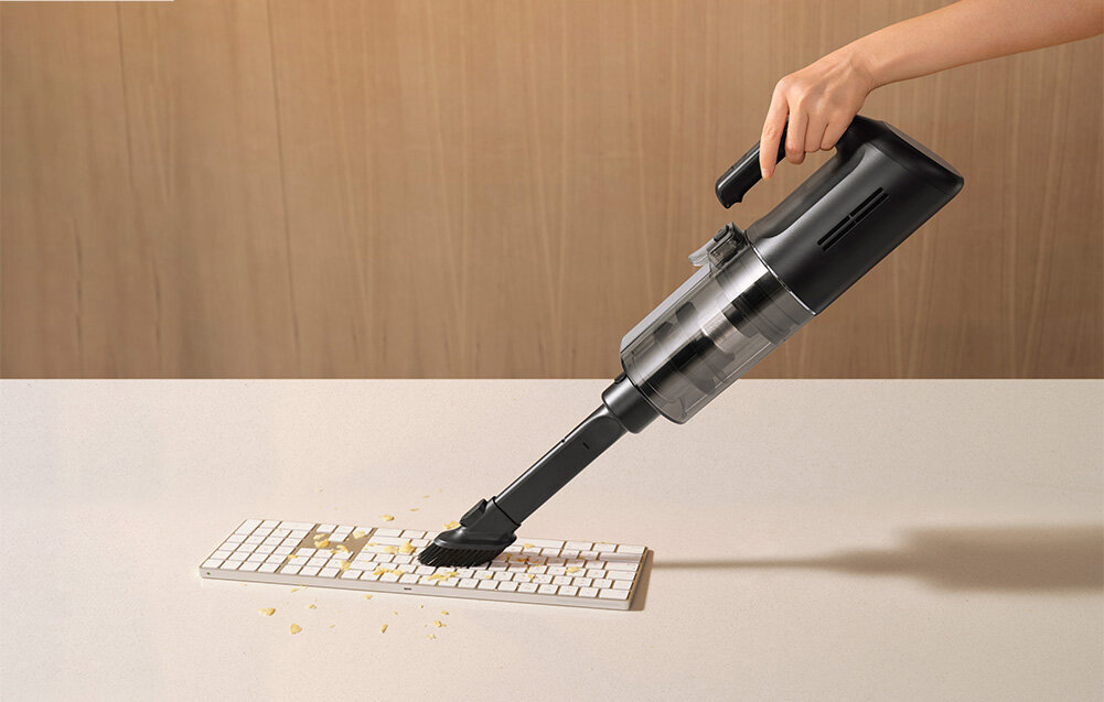 Osoba używa odkurzacza do czyszczenia klawiatury, na której znajdują się okruchy. Odkurzacz jest czarny i ma przezroczysty pojemnik na kurz.