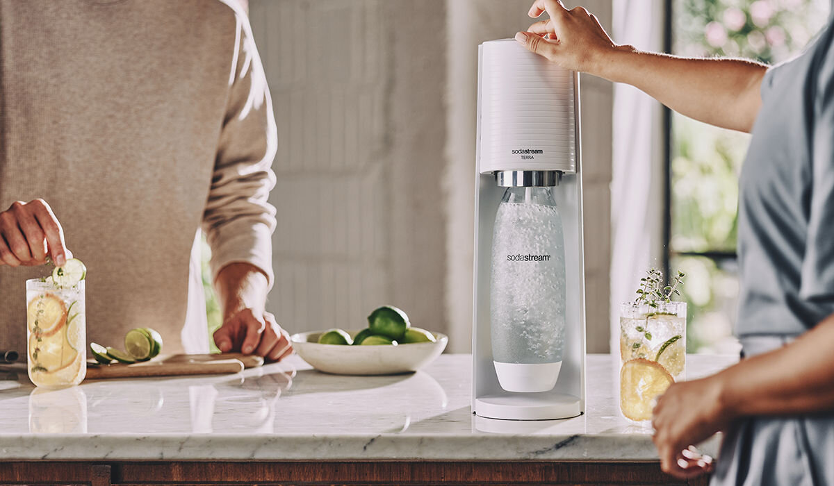 Na blacie kuchennym stoi saturator z butelką wypełnioną wodą gazowaną. Obok znajdują się szklanki z napojem i plasterkami cytryny oraz deska z limonkami.