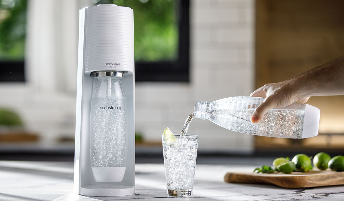 Saturator z logo \'sodastream\' z butelką wypełnioną gazowaną wodą. Obok ręka nalewa wodę z butelki do szklanki z plasterkiem limonki.