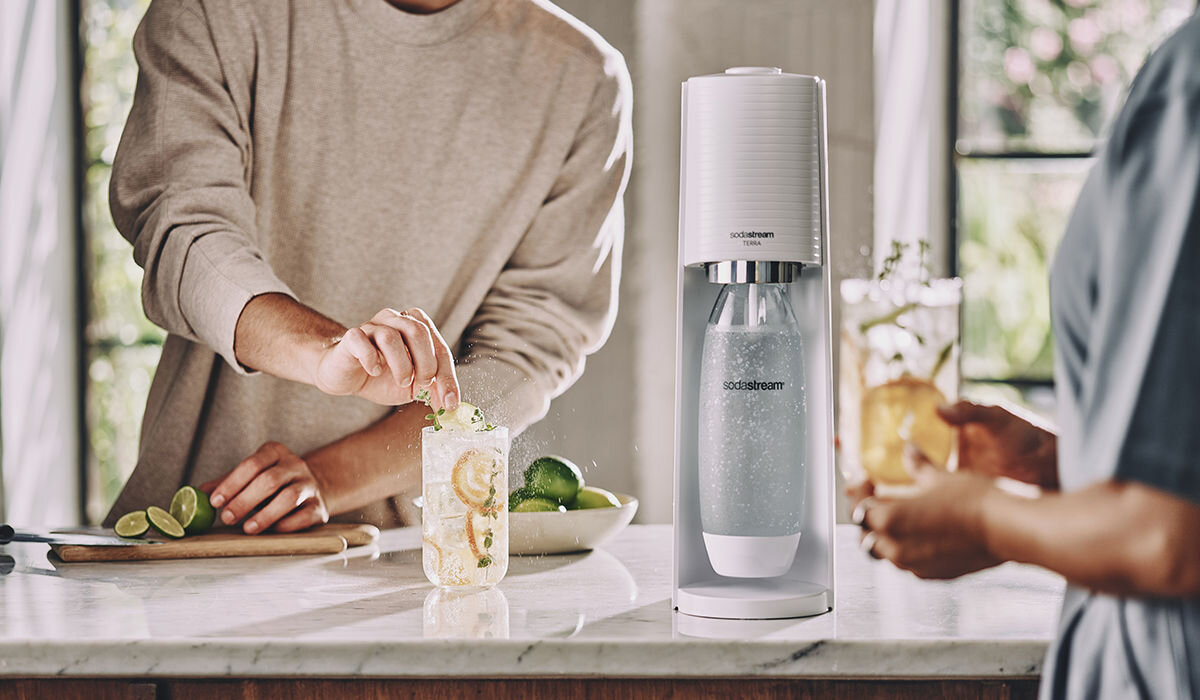 Na blacie kuchennym stoi saturator z widocznym logo \'SODASTREAM\'. Obok znajduje się szklanka z napojem i plasterkami cytryny oraz miska z limonkami.