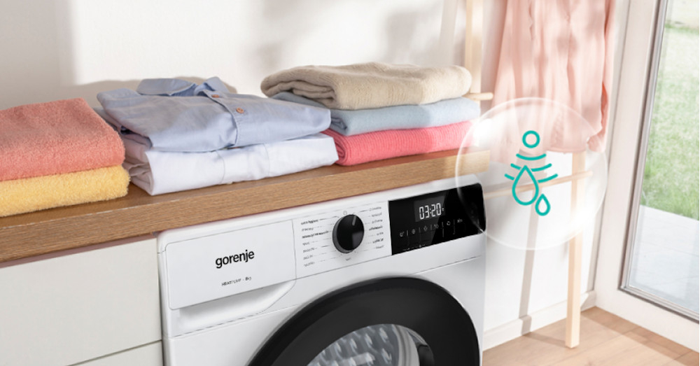 Na obrazie widoczna jest suszarka GORENJE z panelem sterowania, na którym wyświetla się czas 03:20. Na blacie nad suszarką ułożone są poskładane ręczniki i ubrania, a obok znajduje się symbol kropli wody.