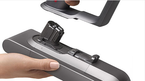 Osoba trzyma szarą część odkurzacza z widocznym miejscem na baterię. Nad nią znajduje się rękojeść z przyciskiem.