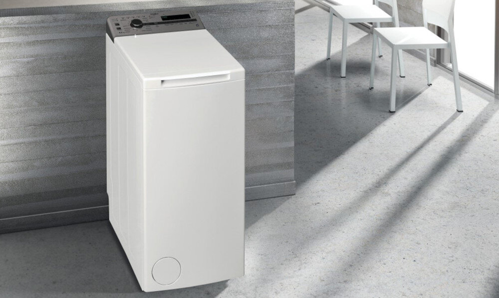Na obrazie znajduje się biała pralka ładowana od góry, ustawiona w nowoczesnym wnętrzu. W tle widoczne są dwa białe krzesła przy metalicznym stole.
