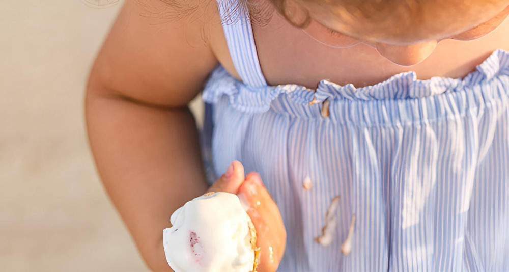 Dziecko w niebieskiej, prążkowanej sukience trzyma loda, który się topi. Na sukience widoczne są plamy od lodów.