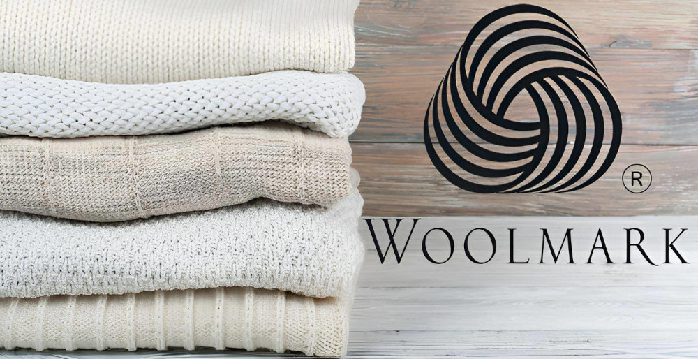 Stos złożonych swetrów w różnych odcieniach beżu i bieli obok logo Woolmark na drewnianym tle.