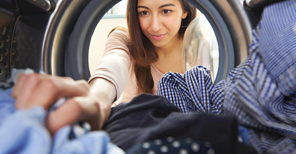 Osoba zagląda do wnętrza pralki, w której znajdują się różnokolorowe ubrania. Widoczna jest metalowa obręcz bębna pralki.