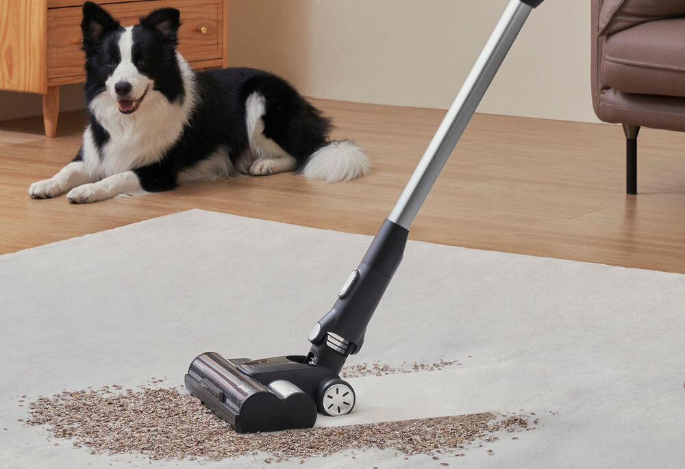 Odkurzacz z aktywnym mopowaniem usuwa rozsypane zanieczyszczenia z dywanu. W tle widoczny jest pies siedzący na podłodze.