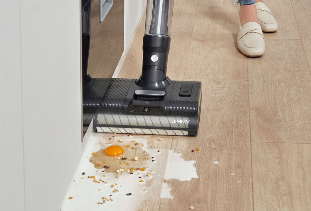 Odkurzacz z logo \'JIMMY\' sprząta rozlane jedzenie na podłodze, w tym jajko i płatki. Obok widoczne są nogi osoby w jasnych butach.