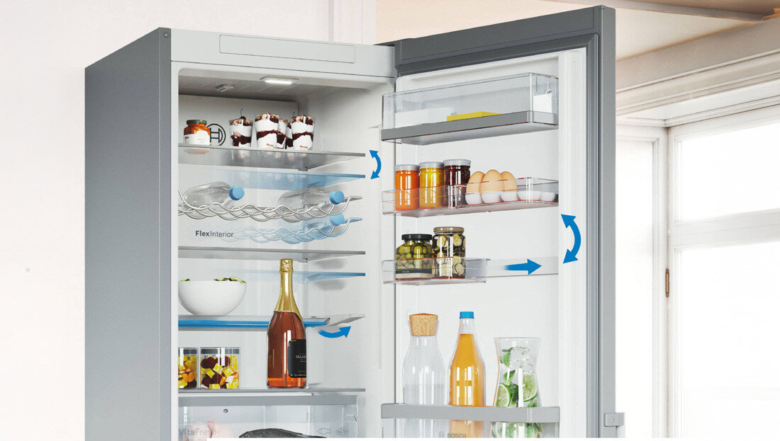 LODÓWKA BOSCH KGN39LBCF BLACK Zdjęcie pokazuje otwartą lodówkę z półkami i pojemnikami, na których umieszczone są różnorodne produkty spożywcze. Niebieskie strzałki ilustrują cyrkulację powietrza oraz funkcje ułatwiające organizację wnętrza Flex Interior składana półka półki na drzwiach