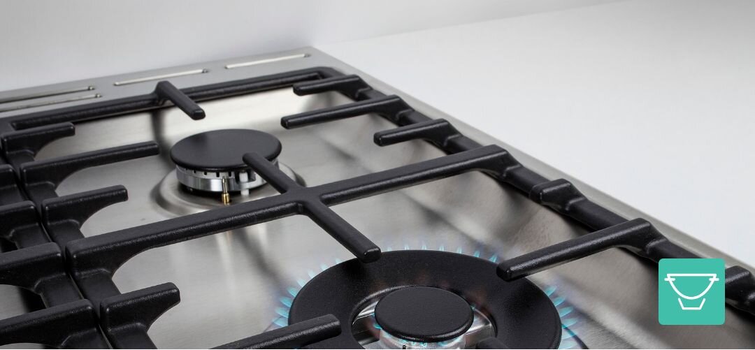 KUCHNIA GORENJE GKS6C71CLI Zbliżenie na płytę gazową z masywnymi, żeliwnymi rusztami na tle stalowej powierzchni, na przednim palniku widoczny jest niebieski płomień, a ikona w rogu sugeruje idealne podparcie garnków i patelni palnik wok mocny płomień azjatyckie dania