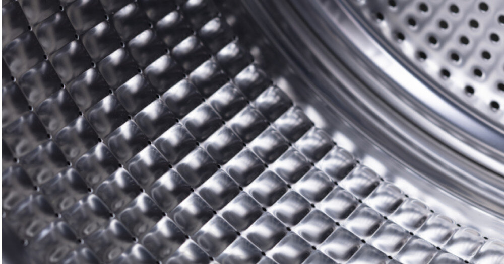 Zbliżenie na wnętrze bębna pralko-suszarki z widoczną metalową powierzchnią o strukturze diamentowej. W tle widać perforowane otwory bębna.