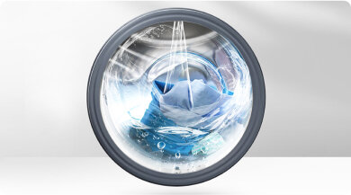 Obraz przedstawia bęben pralki wypełniony wodą i praniem, widoczny przez przezroczyste drzwiczki. Woda tworzy wir, a w środku znajdują się niebieskie tkaniny.