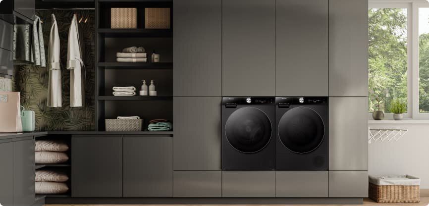 W nowoczesnej pralni znajdują się dwie czarne pralki w zabudowie, otoczone szafkami i półkami z ręcznikami oraz koszami. Przez okno widać zieleń na zewnątrz.