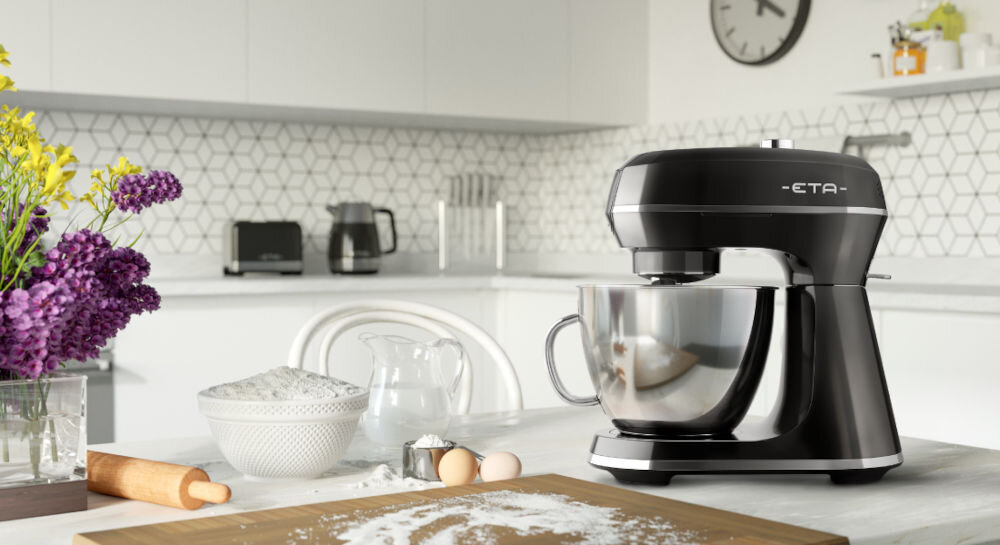 Czarny robot kuchenny planetarny z metalową misą stoi na blacie w nowoczesnej kuchni. Obok znajdują się składniki do pieczenia, takie jak mąka, jajka i wałek do ciasta.
