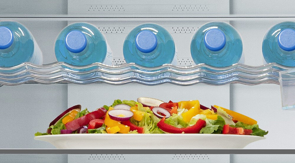 Wnętrze lodówki z półką, na której znajdują się butelki z wodą oraz talerz z sałatką z kolorowych warzyw. Butelki są ułożone poziomo, a sałatka zawiera m.in. paprykę, cebulę i sałatę.
