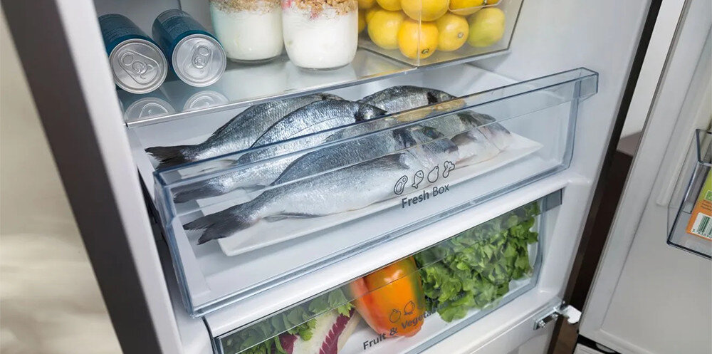 Wnętrze lodówki z widocznymi półkami, na których znajdują się ryby w szufladzie oznaczonej \'Fresh Box\', puszki, słoiki z jogurtem, cytryny oraz warzywa w dolnej szufladzie oznaczonej \'Fruit & Veg\'.