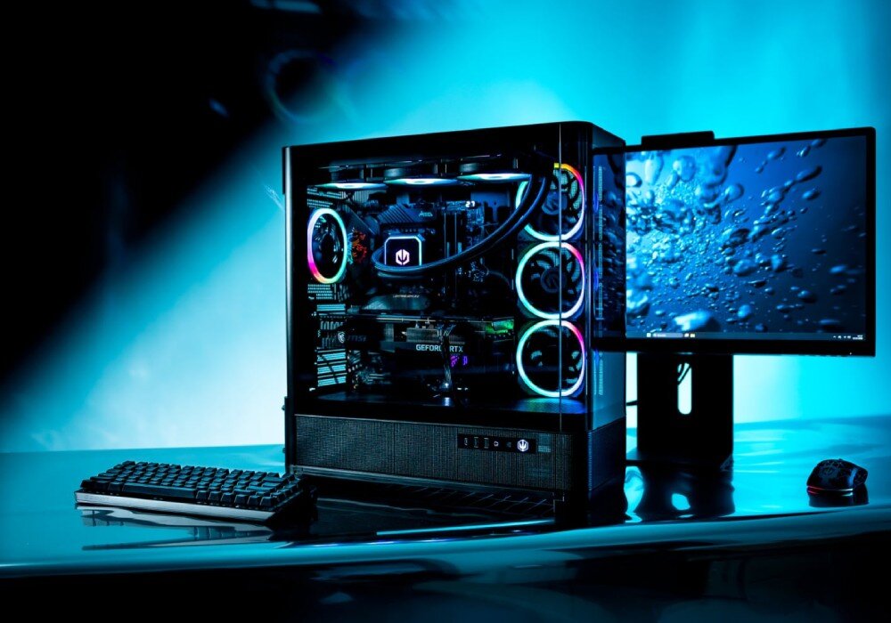Na obrazie widoczna jest obudowa komputera z podświetleniem RGB, obok której znajduje się monitor z wyświetlonym obrazem kropli wody. Na biurku leży klawiatura i myszka, a całość jest oświetlona niebieskim światłem.