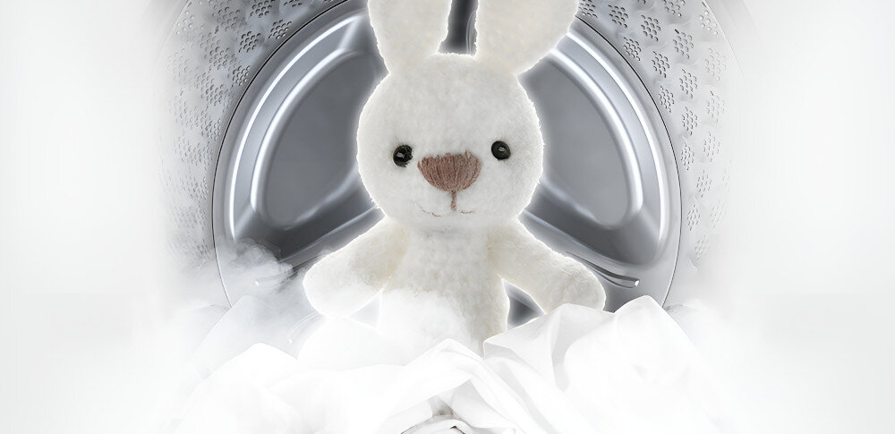 Pluszowy biały królik siedzi w bębnie pralki otoczony białymi tkaninami. W tle widoczna jest metalowa powierzchnia bębna.