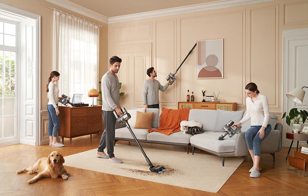 W salonie kilka osób używa odkurzaczy pionowych do sprzątania różnych powierzchni, w tym podłogi i ścian. Na podłodze leży pies, a w tle widać sofę, stolik i dekoracje ścienne.