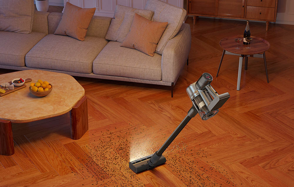 Na drewnianej podłodze w salonie znajduje się odkurzacz pionowy, który zbiera rozsypane drobinki. W tle widać sofę, stolik z owocami i drugi stolik z butelką oraz kieliszkiem.