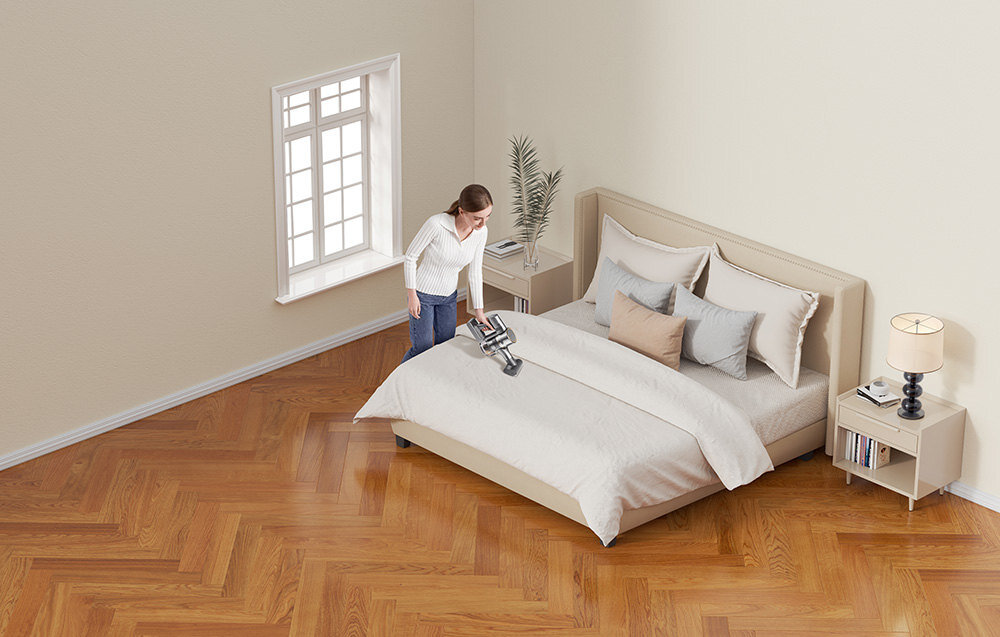 Osoba używa odkurzacza na łóżku w jasnym pokoju z drewnianą podłogą. W pomieszczeniu znajduje się łóżko z poduszkami, stolik nocny z lampką i rośliną oraz okno z białymi ramami.