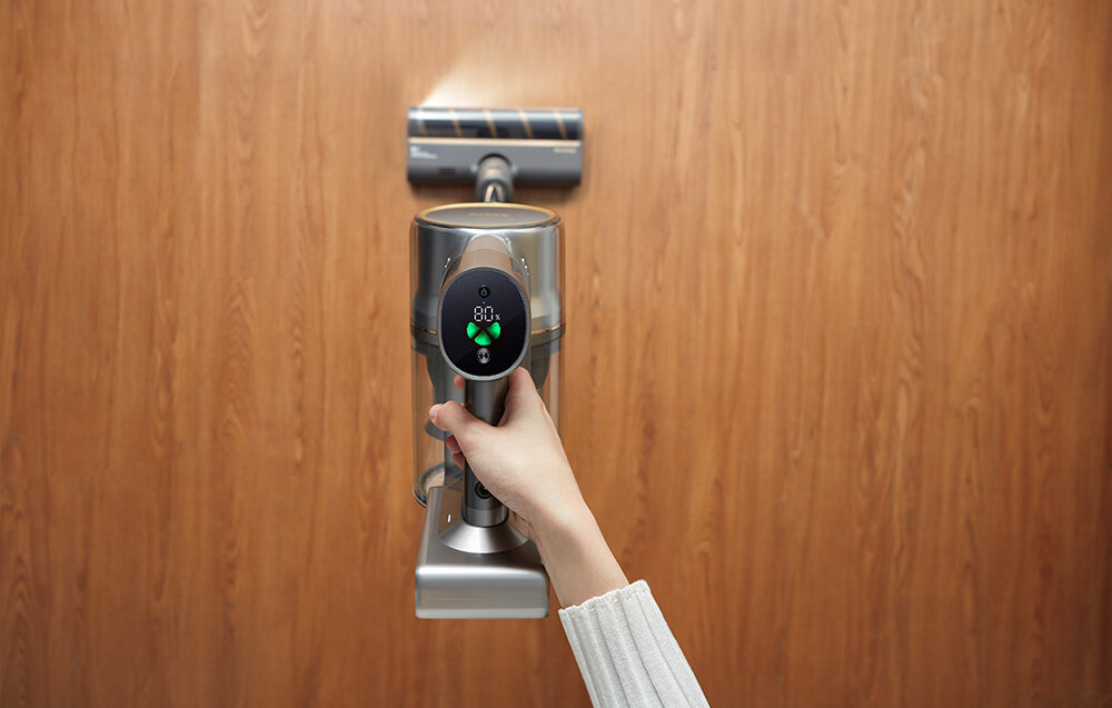 Osoba trzyma odkurzacz z wyświetlaczem pokazującym 80% i ikonami w kształcie strzałek. Odkurzacz jest używany na drewnianej podłodze.