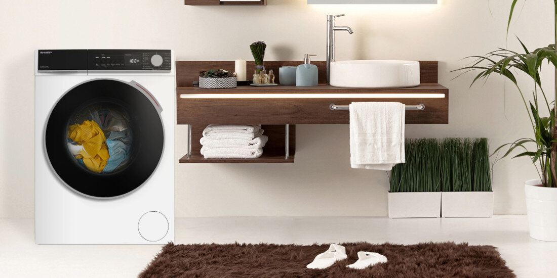 Wnętrze łazienki z pralką SHARP, na której drzwiach widać kolorowe ubrania. Obok znajduje się drewniana szafka z umywalką, ręcznikami i dekoracjami, a na podłodze leży brązowy dywanik.