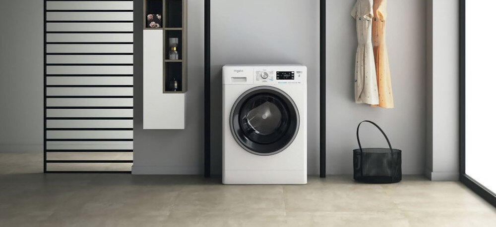 Pralka w białym kolorze stoi w nowoczesnym wnętrzu, obok widać wieszak z ubraniami i półkę z przedmiotami. Na pralce widoczne jest logo \'whirlpool\'.