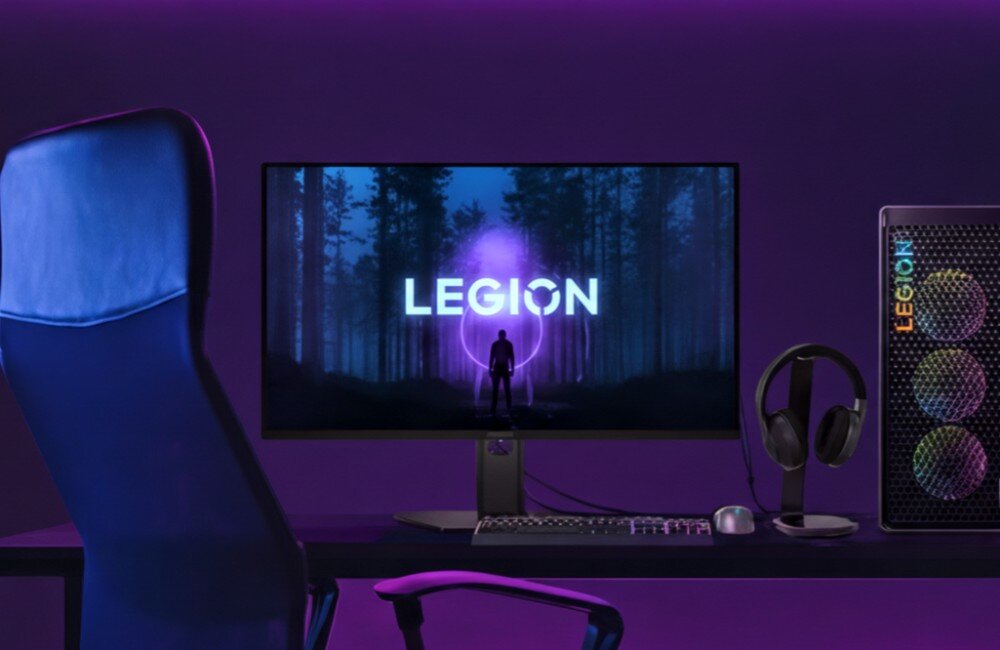 Na biurku znajduje się monitor z wyświetlonym napisem \'LEGION\' na tle lasu oraz komputer z widocznym logo \'LEGION\'. Obok monitora leżą klawiatura, myszka i słuchawki.