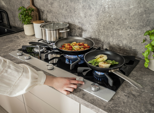 Na płycie gazowej znajdują się trzy garnki z gotującymi się potrawami. Osoba reguluje pokrętło na płycie, a w tle widać kuchenny blat z rośliną i naczyniami.
