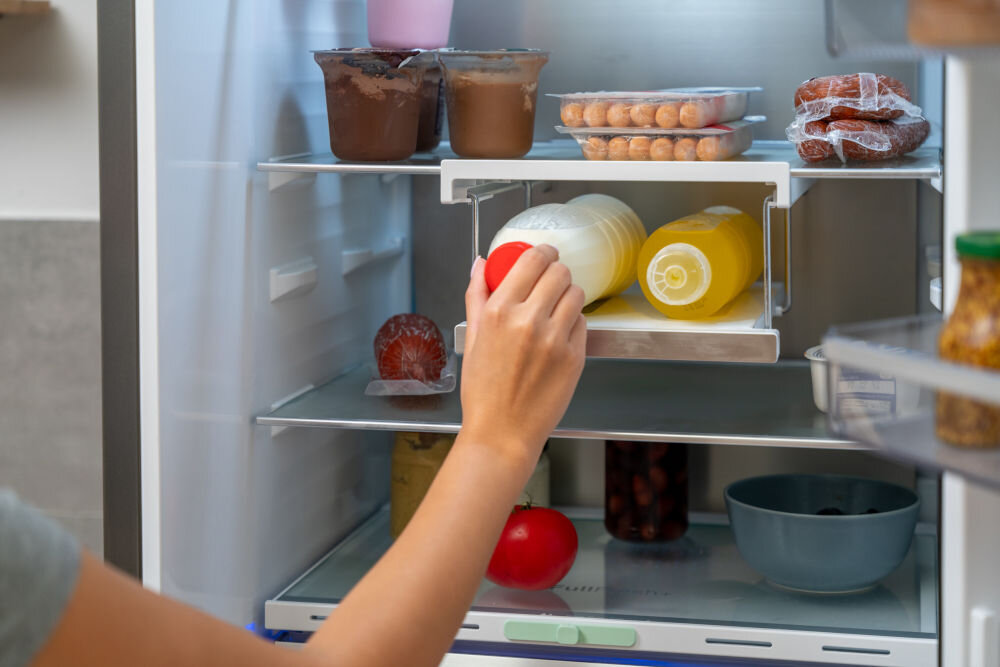 Wnętrze lodówki z różnymi produktami spożywczymi, w tym jogurtami, napojami i warzywami. Osoba sięga po pomidora z półki.