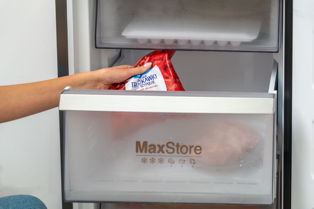 Osoba wkłada opakowanie mrożonych truskawek do szuflady lodówki oznaczonej napisem \'MaxStore\'.