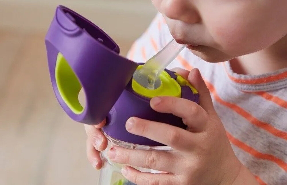 Dziecko pije z fioletowo-zielonej butelki z przezroczystą słomką. Butelka ma uchwyt i otwieraną pokrywkę.