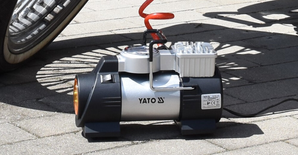 Na obrazie widoczny jest kompresor samochodowy z logo YATO, umieszczony na chodniku. Obok kompresora znajduje się czerwony przewód spiralny, a w tle widać fragment koła pojazdu.