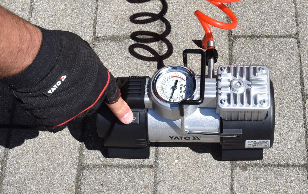 Osoba w rękawicy obsługuje kompresor samochodowy z widocznym manometrem i spiralnym przewodem. Na urządzeniu znajduje się logo \'YATO\'.