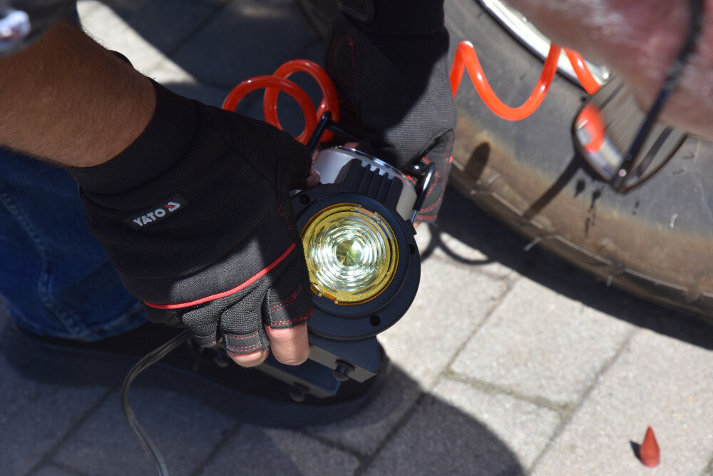 Osoba w rękawiczkach trzyma kompresor samochodowy z widocznym żółtym światłem. Na rękawiczkach widoczny jest napis \'YATO\'.