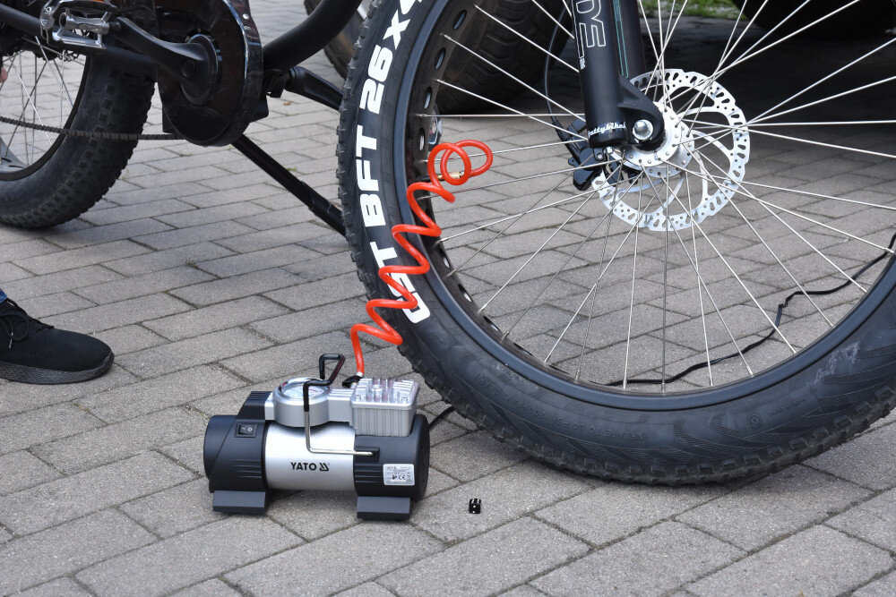 Na obrazie widoczny jest kompresor samochodowy z logo YATO, podłączony do opony rowerowej za pomocą czerwonego węża. Opona ma napis \'BFT 26x4.0\'.
